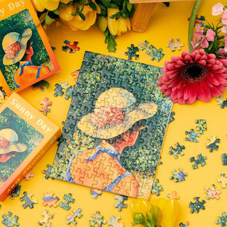 The Minizz Sunny Day 99 Piece Jigsaw Puzzle, featuring a person in a hat, is displayed on a yellow surface with loose pieces, flowers, and the puzzle box nearby.