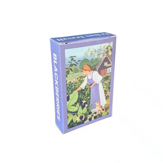 A purple box labeled “Blackberries | 99 Piece Jigsaw Puzzle” by Trevell features Nicolle Lalonde’s illustration of a woman picking blackberries in a country garden, with a cottage in the background—perfect for anyone looking for a charming mini puzzle.