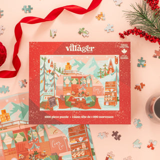 The Villager Puzzles "Camper Cafe" 1,000-piece jigsaw puzzle features a cozy winter campsite illustration on a peach background, with loose pieces, a red ribbon, and holiday greenery adding festive touches.
