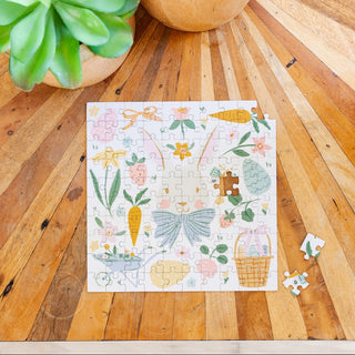 The Easter Bunny | 100 Piece Jigsaw Puzzle by My Mind's Eye, featuring a bunny, carrots, basket, and flowers in pastel colors, sits partially completed on a wooden table beside potted plants.