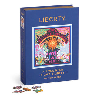 A blue Galison box holds the “All You Need Is Love & Liberty” 500-piece jigsaw puzzle, featuring colorful artwork on recycled paper, with several loose pieces displayed in front.