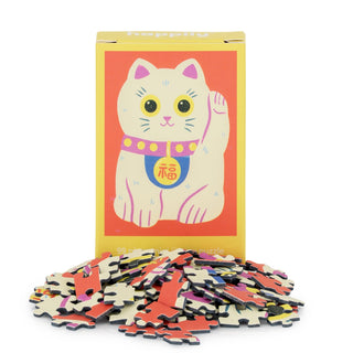 A box of Happily Jigsaw Puzzles’ Lucky Cat | 99 Piece Mini Jigsaw Puzzle, featuring Naomi Wilkinson’s waving cat illustration, stands behind a pile of colorful puzzle pieces.
