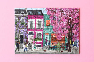 The Notting Hill 99 Piece Jigsaw Puzzle by Piecely features a vibrant street scene with people, a blooming pink tree, plant-adorned buildings, and a pink background—crafted from recycled cardboard.