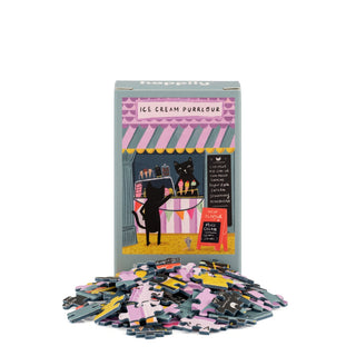 A box labeled Ice Cream Purrlour | 99 Piece Mini Jigsaw Puzzle by Happily Jigsaw Puzzles, featuring a cat selling ice cream, appears behind a small pile of matching puzzle pieces—making this the perfect cat-themed challenge for fans of Nia Gould’s whimsical designs.