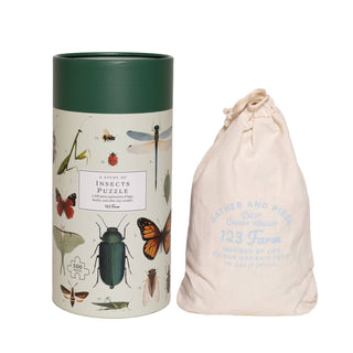 A cylindrical box labeled "Insects | 500 Piece Jigsaw Puzzle" by 123 Farm with insect illustrations sits next to a drawstring bag with blue text.