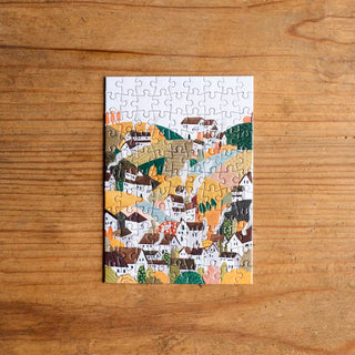 A finished Trevell Autumn Village | 99 Piece Jigsaw Puzzle rests on a wooden surface, highlighting the vibrant and charming autumn village details in this Trevell puzzle.