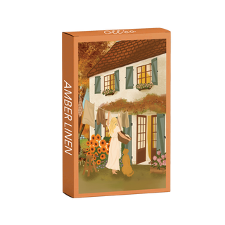 The Amber Linen | 99 Piece Jigsaw Puzzle by Olleo Puzzles features Nicolet Boon's artwork of a woman with a dog hanging laundry outside a flower-adorned cottage with green shutters.