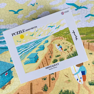 A box of Aparté’s Montalivet | 1,000 Piece Jigsaw Puzzle with a beach scene by Paola Hirou sits atop the finished puzzle, which features blue cabins, a sandy path, and birds in the sky.
