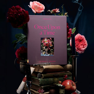 The Once Upon the Time... 1,000 Piece Jigsaw Puzzle by Piecework Puzzles stands upright among pink flowers, candles, and stacked books, with scattered puzzle pieces and smoke curling from a candle nearby.