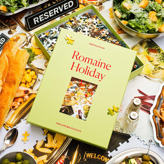 The "Romaine Holiday | 1,000 Piece Jigsaw Puzzle" by Piecework Puzzles sits on a table with salad, bread, olives, scattered pieces, and a Reserved sign in the background.