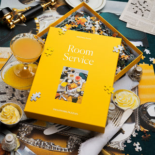 A yellow Room Service | 1,000 Piece Jigsaw Puzzle box by Piecework Puzzles sits on a table with puzzle pieces, a drink, buttered toast, and papers. The set features a playful breakfast in bed theme.