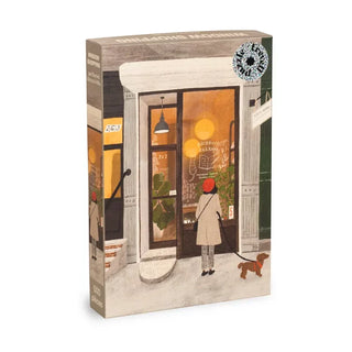 The [Limited Edition] Window Shopping | 500 Piece Jigsaw Puzzle by Trevell showcases a charming illustration by Rachel Victoria Hillis, depicting a person and their small dog outside a warmly lit bookstore with large glass doors revealing potted plants inside.