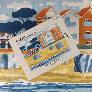 A completed Aparté Soulac sur Mer | 1,000 Piece Jigsaw Puzzle is displayed, with the puzzle box featuring the matching image placed neatly atop the finished seaside scene.