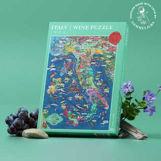 The Water & Wines Italy Wine | 1,000 Piece Jigsaw Puzzle box stands upright on a solid green surface, surrounded by grapes, a plant, stones, and wood—perfect for any aspiring sommelier exploring Italy’s wine regions.