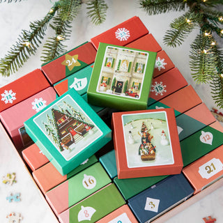 The Janet Hill Advent Calendar by New York Puzzle Company features colorful numbered boxes in a grid, three larger decorated holiday-themed boxes on top, plus pine branches, mini puzzles, and pieces for a festive countdown with 24 100-piece jigsaw puzzles.